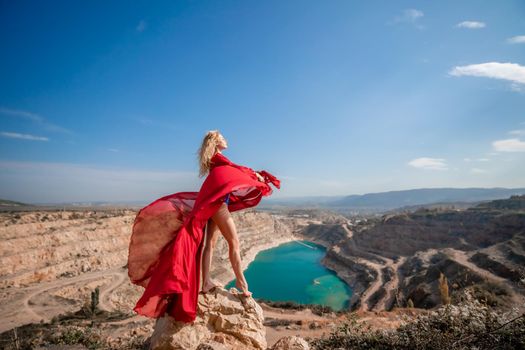 Side view of a beautiful sensual woman in a red long dress posing on a rock high above the lake in the afternoon. Against the background of the blue sky and the lake in the form of a heart.