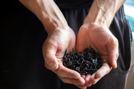 Black small self tapping screws for steel profile of drywall on young adult man hand on light gray table background. Opened palm. Closeup.