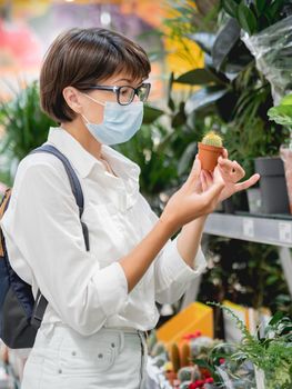 Woman in medical protective mask chooses plants for home. Shelves with seedlings, flowering plants and seeds in flower shop. Indoor agronomic market.