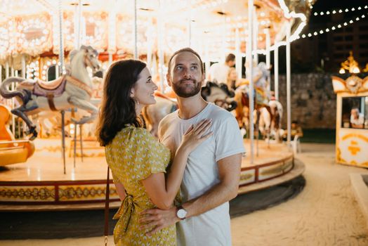 A portrait of a young woman in a yellow dress and her boyfriend with a beard who are staying near the carousel. A couple of lovers on a date at the fair in Valencia.