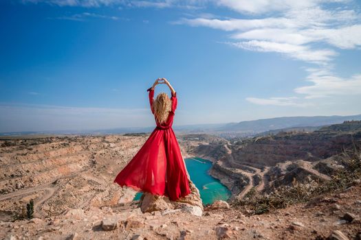 Rear view of a beautiful sensual woman in a red long dress posing on a rock high above the lake in the afternoon. Against the background of the blue sky and the lake in the shape of a heart.