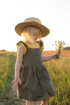 A little blonde girl in a straw hat walks in a field with a bouquet of daisies. The concept of walking in nature, freedom and an eco-friendly lifestyle