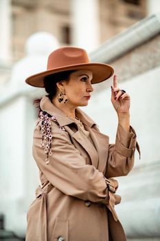 Outdoor fashion portrait of young elegant fashionable brunette woman, model in stylish hat, choker and light raincoat posing at sunset in European city