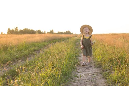 A little blonde girl in a straw hat walks in a field with a bouquet of daisies. The concept of walking in nature, freedom and an eco-friendly lifestyle.