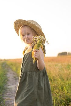 A little blonde girl in a straw hat walks in a field with a bouquet of daisies. The concept of walking in nature, freedom and an eco-friendly lifestyle.