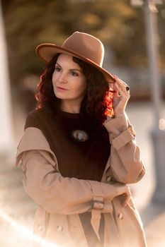 Outdoor fashion portrait of young elegant fashionable brunette woman, model in stylish hat, choker and light raincoat posing at sunset in European city