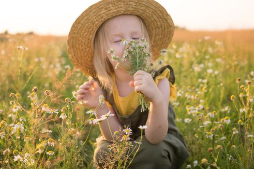 A little blonde girl in a straw hat walks in a field with a bouquet of daisies. The concept of walking in nature, freedom and an eco-friendly lifestyle.