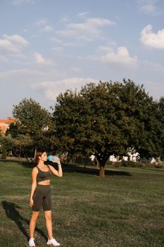 Athletic fit Asian girl drinking water from a bottle in green park. Rest after training