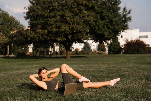 Online training with laptop in the green park. Active fit girl does fitness workout outdoor