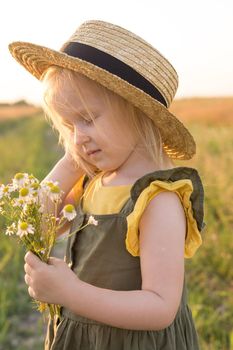 A little blonde girl in a straw hat walks in a field with a bouquet of daisies. The concept of walking in nature, freedom and an eco-friendly lifestyle.