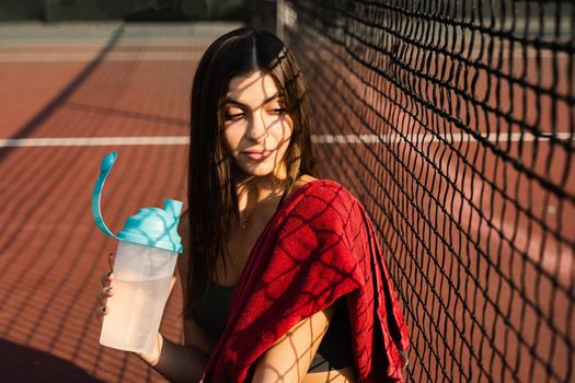 Athletic fit girl drinks water from a bottle. Rest after training on the tennis court