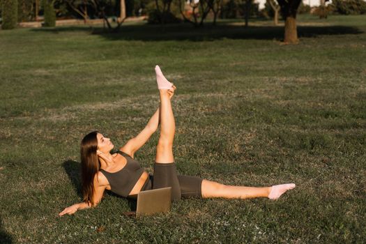 Online training with laptop in the green park. Active fit girl does fitness workout outdoor