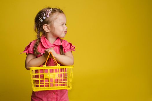 Cunning little cute girl holding small basket posing isolated at yellow studio background. Mischievous charming female child having fun feeling positive emotion medium shot