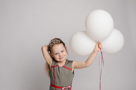 Stock photo of pretty little girl in nice dress and princess crown holding white air balloons and smiling at camera. Studio shot. Isolate on grey background.