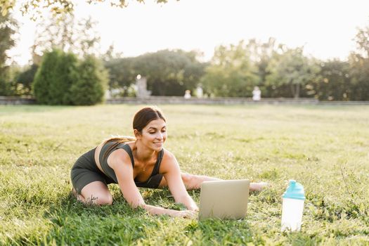 Fit girl with laptop on the green grass. Fitness trainer is chatting online with clients and explain exercises. Sport lifestyle