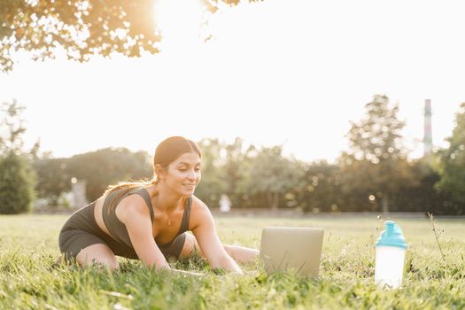 Fit girl with laptop on the green grass. Fitness trainer is chatting online with clients and explain exercises. Sport lifestyle