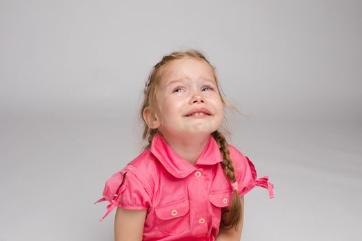 Stock photo of sweet little girl with braids in pink dress crying while sitting on the floor with bare feet. She is looking at the camera while sobbing.