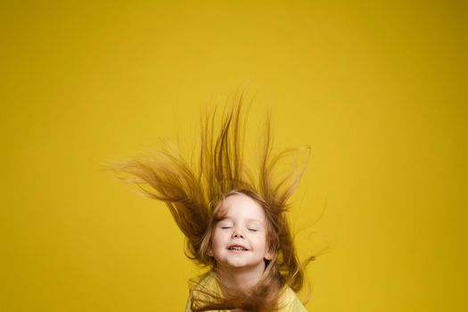 Front view of beautiful longhaired girl in cute shirt playing with hair and twirling in studio. Sunny little child looking at camera and posing on yellow isolated background. Concept of childhood.