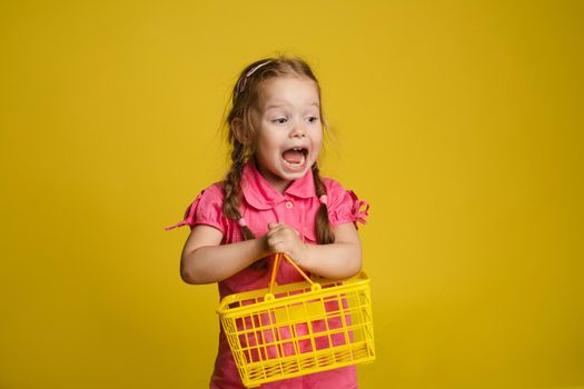 Stock photo portrait of funny lovely little girl in pink dress holding yellow plastic basket and getting excited with her mouth opened. Isolate on yellow background.