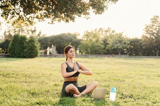 Asian fit girl is sitting in lotus position and meditating online with laptop. Meditation and relaxation outdoors