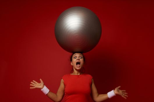 Astonished young sporty woman poses against red wall background with huge fitness ball on her head