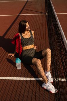 Sport lifestyle. Sport girl with bottle of water and towel. Asian model sitting on the tennis court