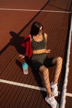 Sport lifestyle. Sport girl with bottle of water and towel. Asian model sitting on the tennis court