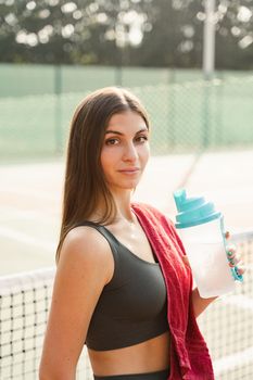 Fit sports model is resting after training and drinking water from a bottle. Sports lifestyle. Beautiful girl posing on the tennis court