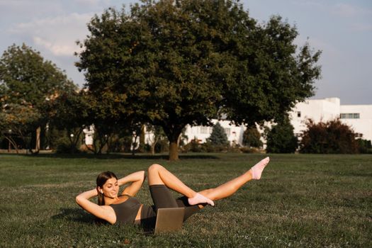 Active fit girl training online with laptop on green grass in the park. Sport lifestyle. Outdoor training