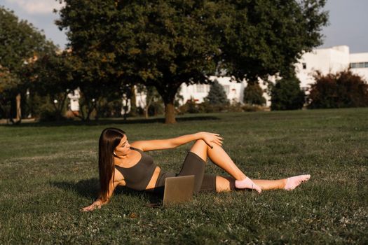 Active fit girl training online with laptop on green grass in the park. Sport lifestyle. Outdoor training