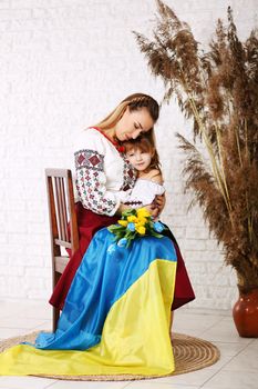 Young beautiful blond woman wearing with little dougter ethnic style embroidered shirt, modern derivative from traditional Ukrainian vyshyvanka with Ukraininan flag
