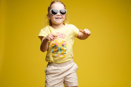 Front view of beautiful longhaired girl in cute shirt playing with hair and twirling in studio. Sunny little child looking at camera and posing on yellow isolated background. Concept of childhood.