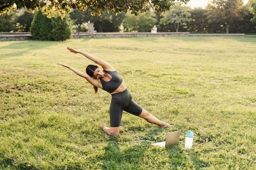 Yoga meditation online with laptop on the grass in green park. Fit girl training outdoor. Relaxation and meditating