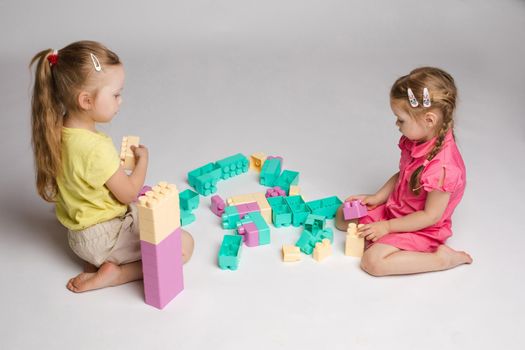 View from side of two cute girls sitting on floor and playing with building blocks in studio. Little children enjoying game, talking and smiling on isolated background. Concept of joy and activity.