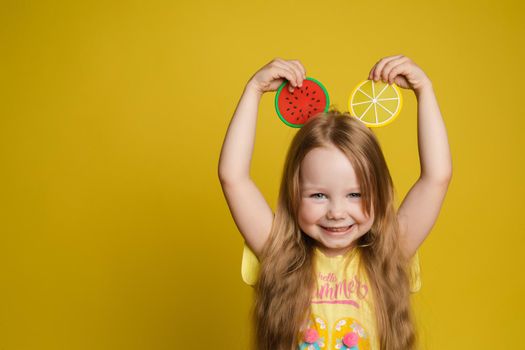 Happy cute little child girl holding slice of fruit over head making ears having fun medium close-up. Smiling beautiful female kid feeling positive emotion posing isolated at yellow studio background