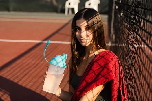 Athletic fit girl drinks water from a bottle. Rest after training on the tennis court