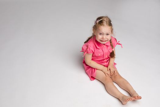 Stock photo of sweet little girl with braids in pink dress crying while sitting on the floor with bare feet. She is looking at the camera while sobbing.