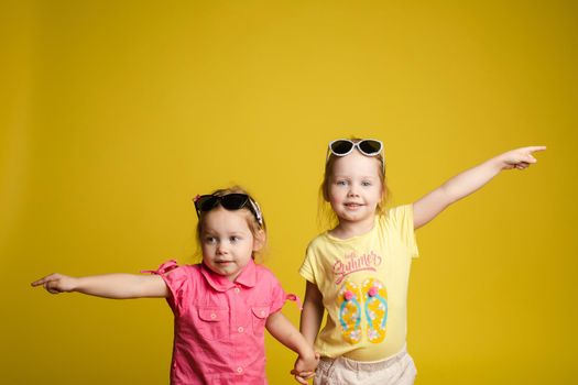 Two happy beautiful stylish little girl wearing sunglasses posing isolated at yellow studio background medium shot. Adorable laughing female child friend enjoying friendship having positive emotion