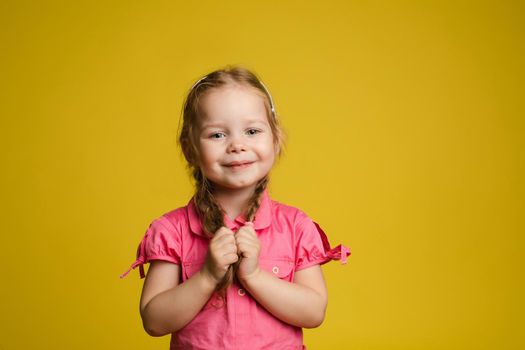 Portrait of adorable little cute female child holding hair posing looking at camera medium shot. Smiling pleasant happy girl having positive emotion isolated at yellow studio background