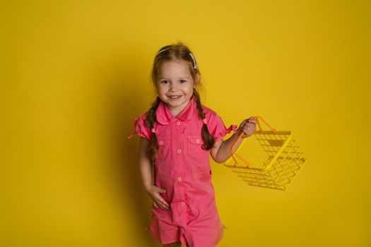 Stock photo portrait of funny lovely little girl in pink dress holding yellow plastic basket and getting excited with her mouth opened. Isolate on yellow background.