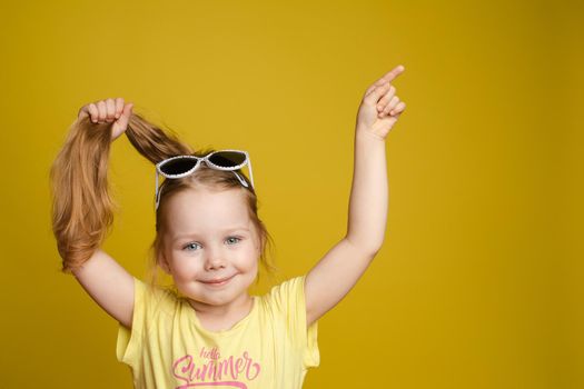 Front view of sunny little girl keeping hair in hand, and pointing with finger at copy space on yellow isolated background. Beautiful child with glasses looking at camera, smiling and posing.