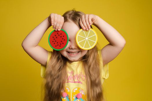 Front view of happy girl standing on yellow isolated background and closing eyes with toys. Cheerful longhaired child laughing and playing hide and seek. Concept of game and happiness.