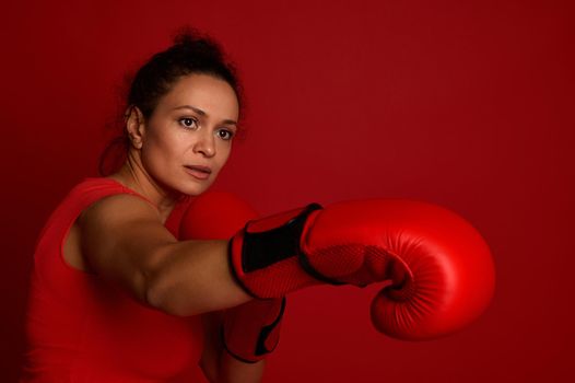 Determined boxer female athlete fights in red boxing gloves on a red background. Martial art concept with copy space for advertisement for boxing day event