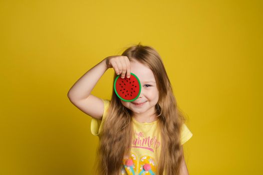 Portrait of smiling cute little girl holding slice of lemon near eye looking at camera medium close-up. Happy female beautiful child having fun posing isolated at yellow studio background