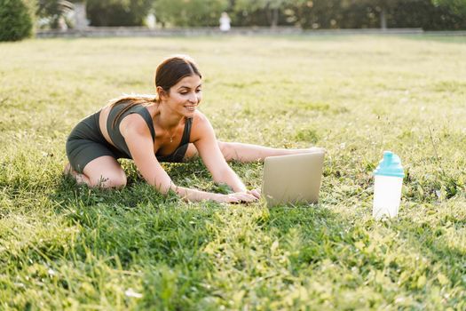 Fit girl with laptop on the green grass. Fitness trainer is chatting online with clients and explain exercises. Sport lifestyle