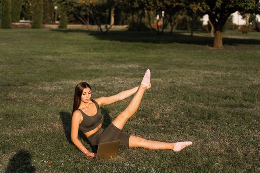 Active fit girl training online with laptop on green grass in the park. Sport lifestyle. Outdoor training