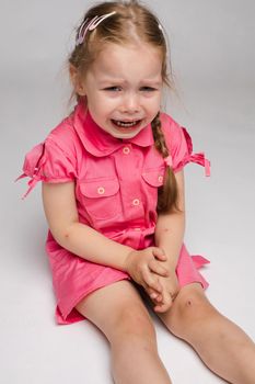 Stock photo of sweet little girl with braids in pink dress crying while sitting on the floor with bare feet. She is looking at the camera while sobbing.