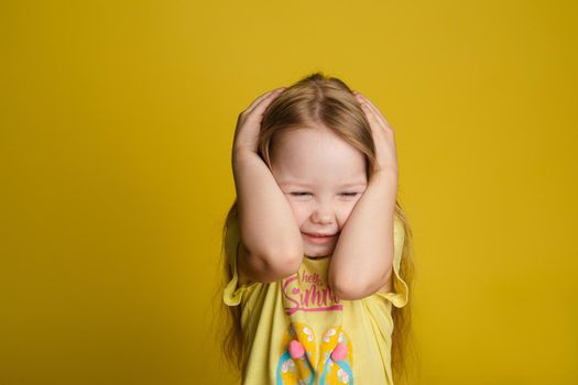 Portrait of scared little girl closing head by hands posing isolated at yellow studio background medium close-up. Cute baby female child holding hand on ears having negative emotion feeling noise