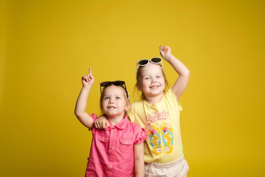 Two happy beautiful stylish little girl wearing sunglasses posing isolated at yellow studio background medium shot. Adorable laughing female child friend enjoying friendship having positive emotion