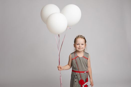 Stock photo of pretty little girl in nice dress and princess crown holding white air balloons and smiling at camera. Studio shot. Isolate on grey background.